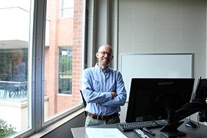 Man smiling with glasses on standing behind a desk.